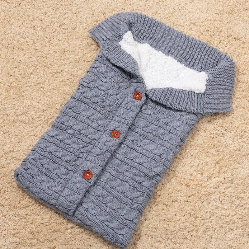 Gray knitted baby sleeping bag with white fleece inside lining and brown buttons with a ribbed neckline.