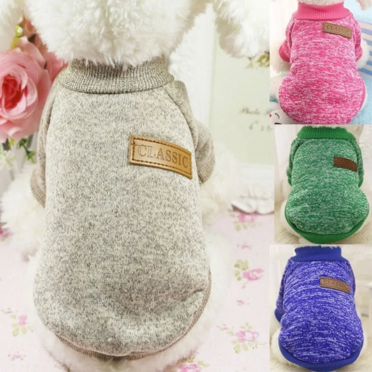 A photo of a stuffed animal wearing a beige and gray sweater with ribbed neck and vegan leather badge that says: Classic. Also seen in pink, green, and indigo.